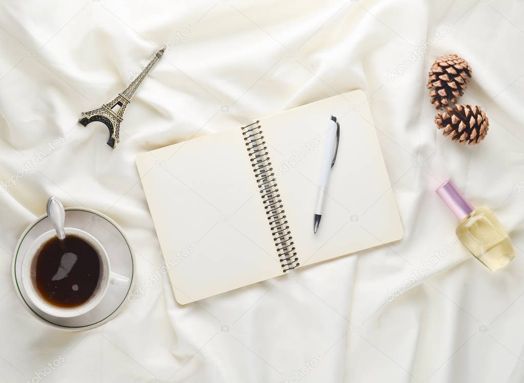 Morning inspiration and desire to trip to Paris. A cup of tea, notebook with a pen, pine cone, souvenir statue, perfume on a white bed sheet. Breakfast on the bed. Top view.