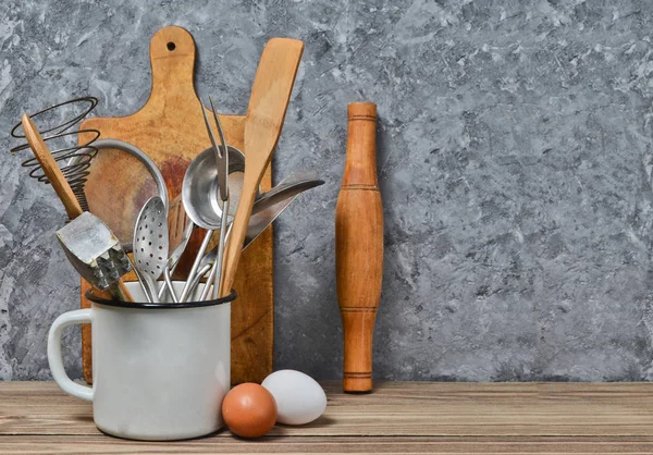 Ferramentas de cozinha para cozinhar em uma mesa de madeira no fundo de uma parede de concreto. Colher, garfos, espátula de madeira, rolo, ovos . — Fotografia de Stock