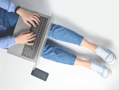 Blogging, bir dizüstü bilgisayarda çalışma kavramı. Beyaz bir katta oturan bir dizüstü bilgisayar kullanarak. Modern teknolojiler. Kot pantolon ve spor ayakkabı kadın bacaklar. Üstten Görünüm. Düz yatıyordu.
