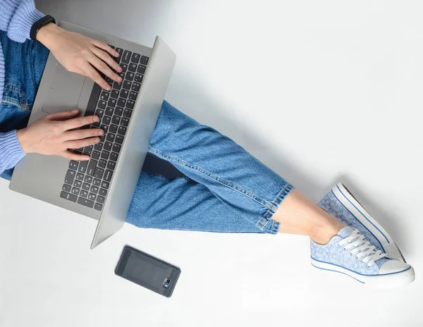 Blogging, bir dizüstü bilgisayarda çalışma kavramı. Beyaz bir katta oturan bir dizüstü bilgisayar kullanarak. Modern teknolojiler. Kot pantolon ve spor ayakkabı kadın bacaklar. Üstten Görünüm. Düz yatıyordu.
