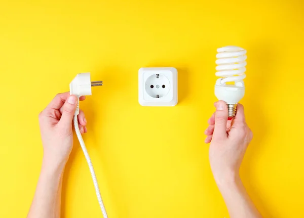 Female hands plug the power plug into electrical outlet while holding spiral light bulb on yellow background. Minimalism