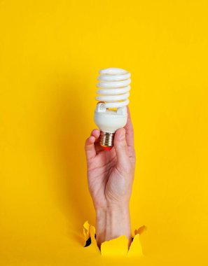 Kadın eli, sarmal ampulü yırtılmış sarı kağıdın içinden tutuyor. Minimalist yaratıcı moda konsepti