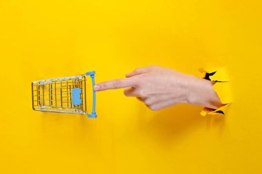 Kadın el, yırtık sarı kağıdın içinden geçen küçük bir alışveriş arabasına dokunuyor. Asgari alışveriş konsepti