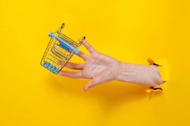 Dişi el, yırtık sarı kağıtlar arasında mini alışveriş arabası tutuyor. Asgari alışveriş konsepti