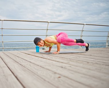 Spor giyim ve şapkalı genç bir kadın sabahları plajda yoga egzersizi yapıyor.