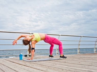 Spor giyim ve şapkalı genç bir kadın sabahları plajda yoga egzersizi yapıyor.