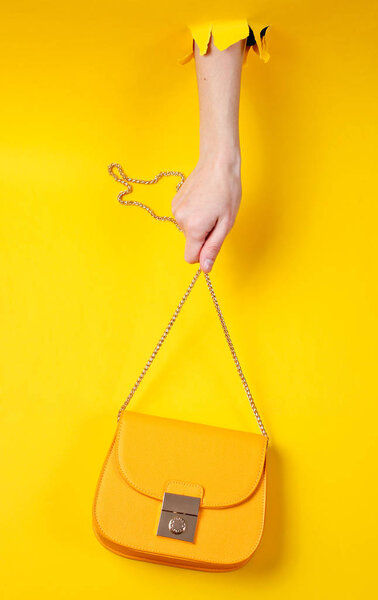 Female hand is holding yellow leather bag over chain through torn yellow paper. Minimalistic fashion concept