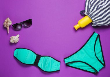 Mor arka planda yaz plaj kıyafetleri ve aksesuarları. Kadın mayosu, güneş gözlüğü, plaj çantası. Üst görünüm
