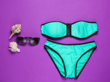 Mor arka planda yaz plaj kıyafetleri ve aksesuarları. Kadın mayosu, güneş gözlüğü. Üst görünüm