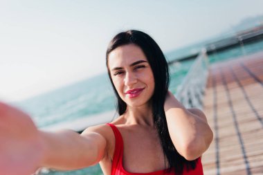 Kırmızı mayo giymiş genç bir kadının portresi sahilde selfie çekiyor.