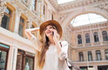 Yaşlı şehir mimarisinin arka planında konuşan keçeli şapkalı neşeli sarışın kadın.