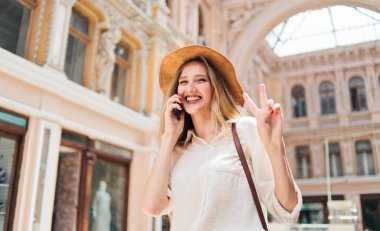 Telefondan konuşan keçeli şapkalı neşeli sarışın kadın ve eski şehir mimarisinin arka planındaki kameraya bak.