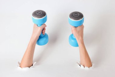 Beyaz arka plandaki yırtık deliklerden bir dambılı tutan kadın eller. Yaratıcı spor sanatı