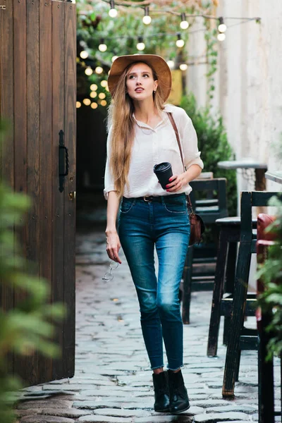 Young attractive woman in casual style clothes walking along the courtyard of the old city with a cup of coffee on the go