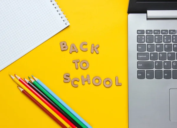 Back to school minimalistic concept. Laptop, color pencils, notebook on yellow background. Slogan Back to school with wooden letters