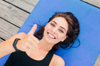 Gülümseyen sporcu kadın mindere uzanıyor ve baş parmağını kaldırıyor. Üst görünüm portresi