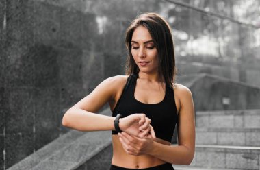 Dışarıdaki gri mermer merdivenlerin arka planında akıllı bir bileziğe bakan genç bir kadın. Spor Malzemeleri