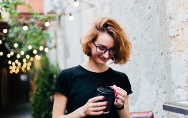 Cheerful hipster girl in glasses with short haircuts holding coffee cup against the background of street garlands outdoor
