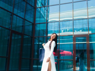 Shopping concept. Afro woman shopaholic in white beautiful dress holding many paper shopping bags in front of business building with blue windows