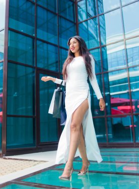 Cheerful Afro woman in white beautiful dress holding many paper shopping bags while walking  at front of business building with blue windows. Shopaholic concept. 