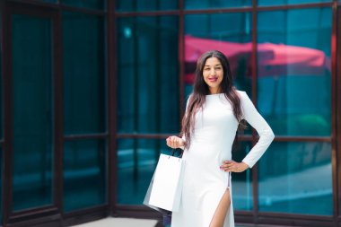 Beauty afro woman in white beautiful dress holding paper shopping bags on background of business building with blue windows