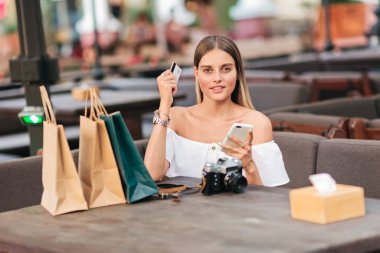İnternetten alışveriş. Alışveriş meraklısı genç bir kadın alışveriş çantalarıyla masada oturuyor ve elinde akıllı telefon ve açık kafedeki banka kartını tutuyor.