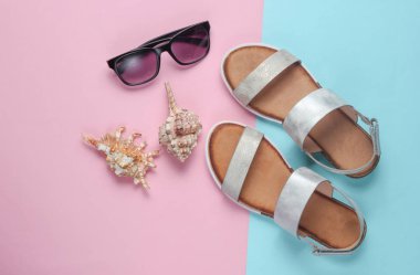 Yaratıcı yaz plajı düzlüğü. Deri kadın sandaletleri, deniz kabukları, pastel mavi-pembe arka planda güneş gözlüğü. Üst görünüm