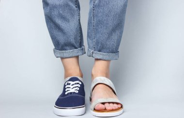 Kot pantolonlu kadın bacakları spor ayakkabısı ve beyaz arka planda sandalet. Mevsimlik ayakkabılar. Yaratıcı moda çekimi