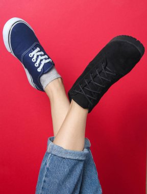 Pantolonlu, çizmeli ve kırmızı arka planda spor ayakkabılı kadın bacakları. Mevsimlik ayakkabılar. Yaratıcı moda çekimi