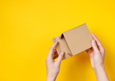 Kadın elleri sarı arka planda boş karton kutuyu açar. Üst görünüm
