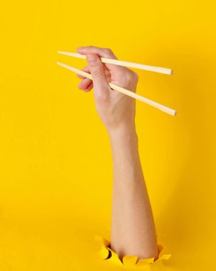 Kadın yemek çubuklarını sarı arka plandaki yırtık delikten tutuyor. Minimalist gıda konsepti. Üst görünüm