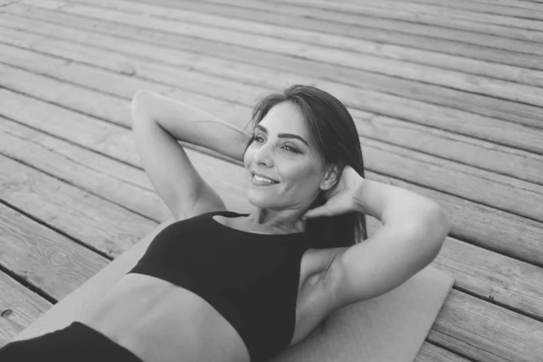 木製の板の上のマットの上に横たわる腹部の筋肉のための練習をする美しいスポーツの女性屋外 黒と白 — ストック写真