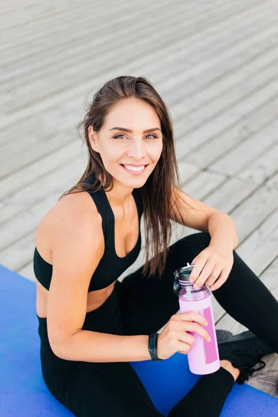 Healthy fit woman sits on a gymnastic mat and holds bottle of water on wooden boards outdoors