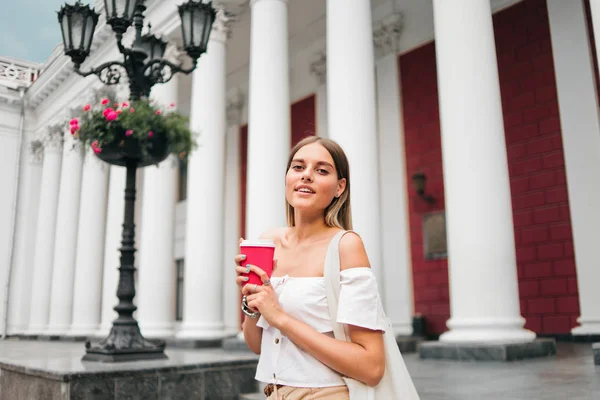 Young fashionable woman holding a cup of coffee on the go against the background of urban architecture with columns