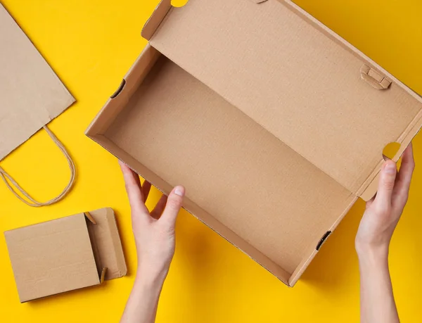 女性的手把一个空的纸板箱放在黄色的背景上 上面有一个纸袋和一个盒子 顶部视图 — 图库照片