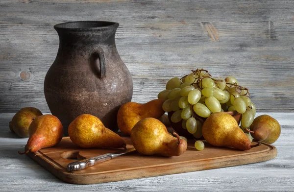 Νεκρή ζωή σε ένα ρουστίκ στυλ: ένα σύνολο από πήλινα πιάτα, σταφύλια και αχλάδια σε ένα ξύλινο τραπέζι. Φυσικό φως από τα παράθυρα. — Φωτογραφία Αρχείου