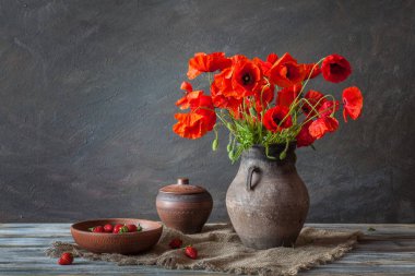 Eski bir çömlek, bir buket kırmızı haşhaş ve ahşap bir masada çilek.