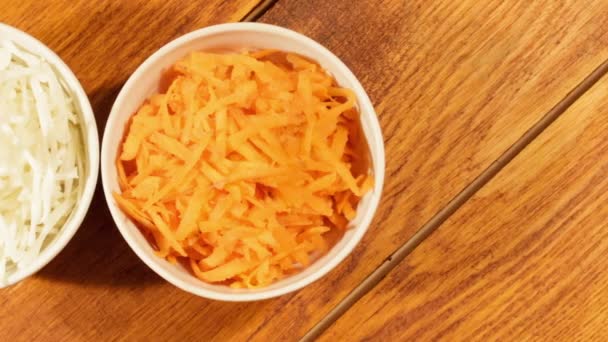 Нарізана морква і капуста в білій чашці на дерев'яному фоні крупним планом. Інгредієнти для овочевого салату — стокове відео