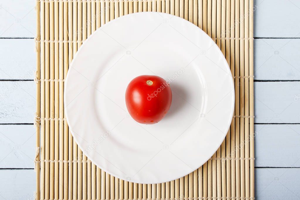 Red ripe tomatoes on white plate. Bamboo mat on wooden table. Top view.