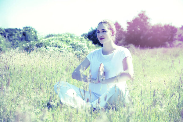 Healthy beautiful yoga girl relaxing and meditating sitting with praying hands for energy exercise in green meadows, contrast, colored and toned effects