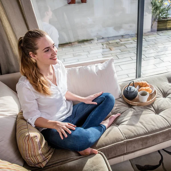 Sorrindo linda jovem sentada no sofá confortável com café da manhã — Fotografia de Stock