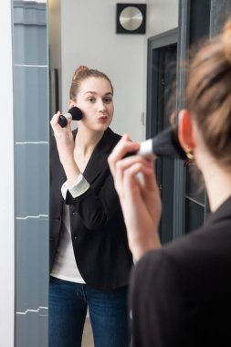 Güzel genç bir kadın aynanın önünde yüzünü pudralıyor.