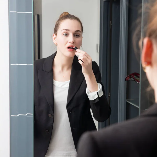 Mujer joven con traje de negocios aplicando su lápiz labial ante el espejo — Foto de Stock