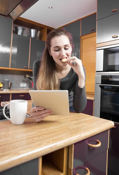 Chica adolescente conectada leyendo tableta en la cocina, comiendo un cooki — Foto de Stock