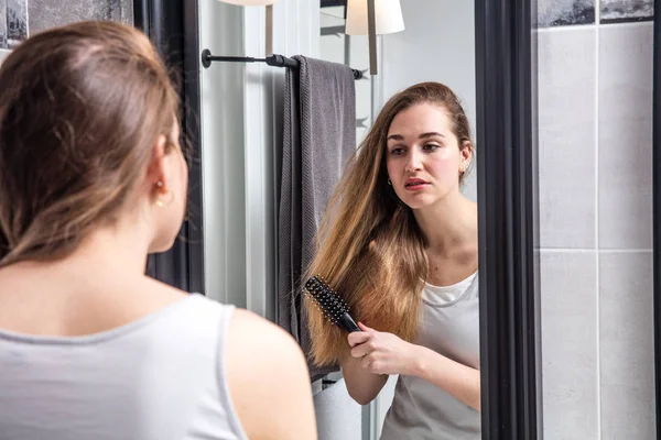Chica pensando cepillarse el pelo largo de pie delante del espejo — Foto de Stock