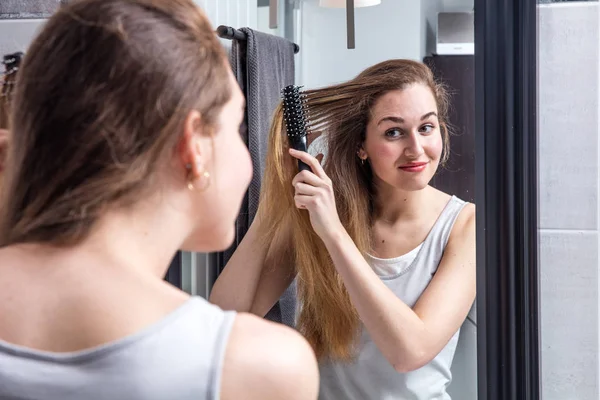 Feliz joven mujer cepillando el pelo largo delante del espejo — Foto de Stock