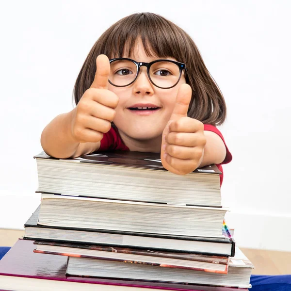 Feliz niño inteligente con los pulgares hacia arriba apoyándose en los libros, interior —  Fotos de Stock