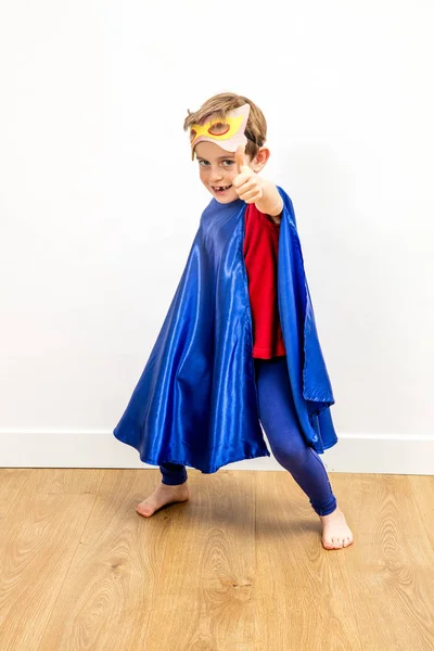fun superhero boy with thumbs up and a mask playing, having super dynamic power over wooden floor for freedom and childhood concept