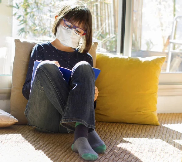 relaxed lockdown kid with healthy face mask writing on exercise notebook sitting for school shutdown, sunny green balcony background