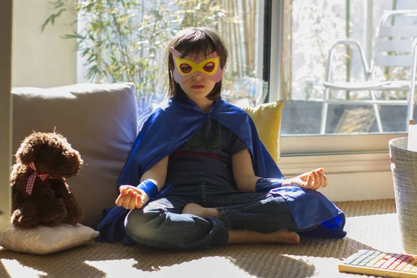 relaxed superhero child dressed up with mask and cape seeking for calm from yoga, meditation and mindfulness facing corona virus lockdown